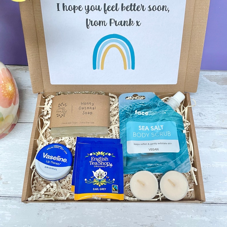 May include: A gift box filled with self-care items, including a bar of honey oatmeal soap, a tube of sea salt body scrub, a small tin of lip balm, two tea bags, and two tea lights. The box is decorated with shredded paper and a card that reads "I hope you feel better soon, from Frank x".