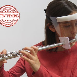 May include: A person wearing a clear plastic face shield with a white foam headband plays a silver flute. The face shield has a white fabric covering the mouthpiece of the flute. A red stamp with the text "PATENT PENDING" is in the upper left corner of the image.