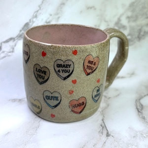 Può includere: Una tazza in ceramica con esterno marrone chiaro e interno rosa. La tazza è decorata con caramelle a forma di cuore con frasi come "Love You" e "Cute". Piccoli cuori rossi sono sparsi intorno alla tazza.