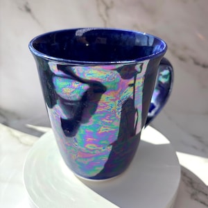 Peut inclure: Une tasse en céramique avec une couleur de base bleu profond et une finition irisée hautement réfléchissante qui affiche des teintes arc-en-ciel changeantes sur sa surface. La tasse présente une large ouverture arrondie et une anse incurvée.