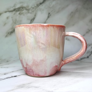 Peut inclure: Une tasse en céramique avec un corps arrondi et une anse. La tasse présente un dégradé de couleurs, passant d'un bord rose rougeâtre à une base blanc crème. La tasse mesure environ 10 cm de haut.