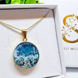 May include: A gold chain necklace with a round pendant featuring a blue and white resin ocean wave design. The pendant is filled with small white and blue pieces of sea glass and sand.