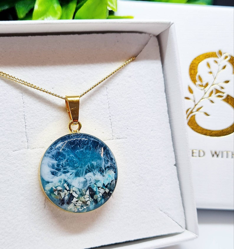 May include: A gold chain necklace with a round pendant featuring a blue and white resin ocean wave design. The pendant is filled with small white and blue pieces of sea glass and sand.