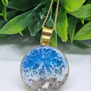 May include: A gold chain necklace with a round pendant featuring a blue and white resin ocean scene with sand at the bottom.