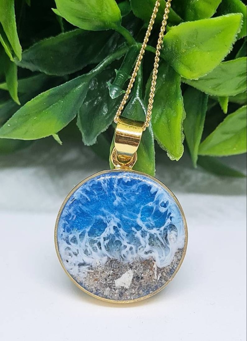 May include: A gold chain necklace with a round pendant featuring a blue and white resin ocean scene with sand at the bottom.