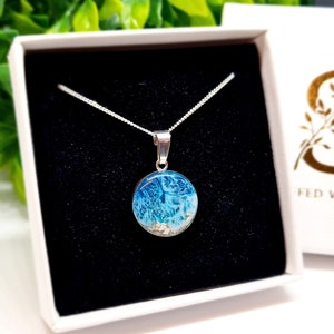 Pu&ograve; includere: Una collana con catena d'argento con un ciondolo rotondo che presenta un design in resina blu e bianca con glitter bianco. Il ciondolo &egrave; in una scatola regalo bianca.