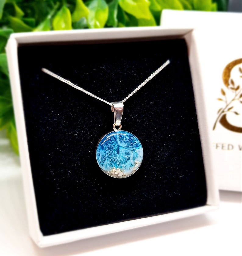Pu&ograve; includere: Una collana con catena d'argento con un ciondolo rotondo che presenta un design in resina blu e bianca con glitter bianco. Il ciondolo &egrave; in una scatola regalo bianca.