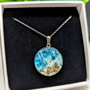 May include: A silver chain necklace with a round pendant featuring a blue and white resin ocean scene with sand and seashells.