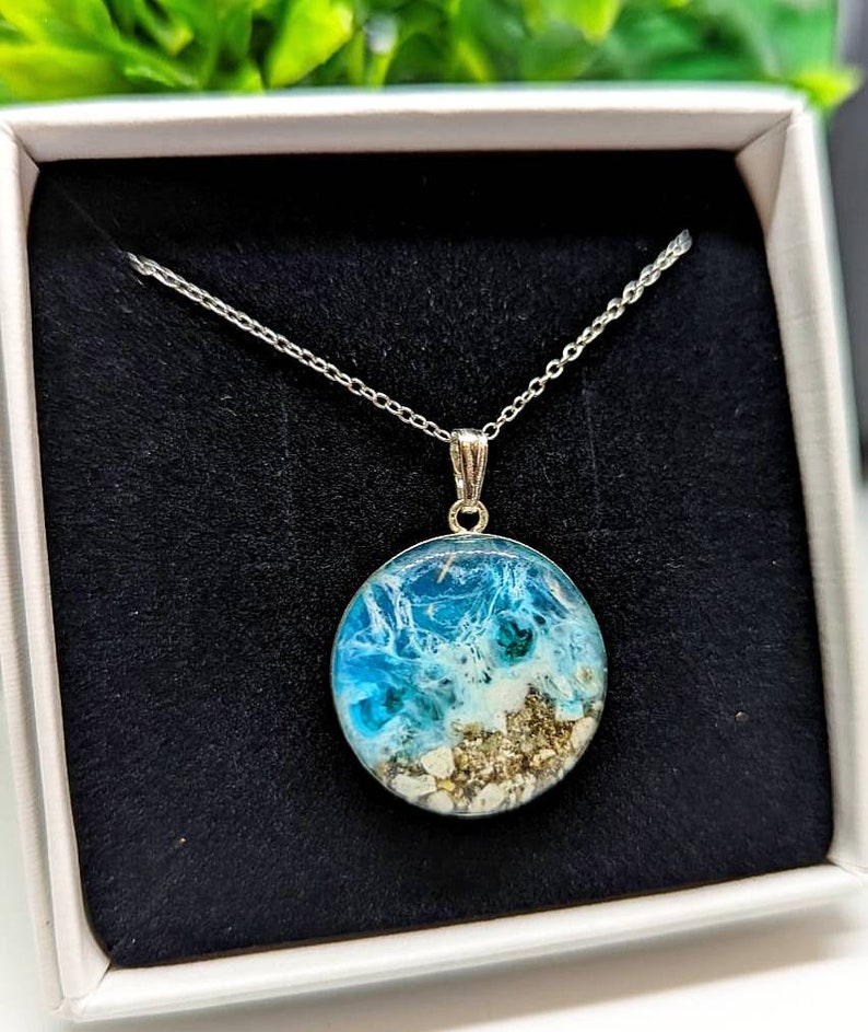 May include: A silver chain necklace with a round pendant featuring a blue and white resin ocean scene with sand and seashells.