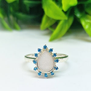 May include: A silver ring featuring a white teardrop-shaped stone encircled by a halo of blue gemstones.