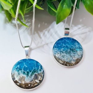 May include: Two silver necklaces with round pendants featuring ocean scenes. The pendants are filled with blue, white, and brown resin, resembling waves crashing on a sandy beach.