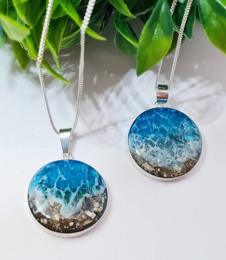 May include: Two silver necklaces with round pendants featuring ocean scenes. The pendants are filled with blue, white, and brown resin, resembling waves crashing on a sandy beach.