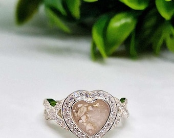 Può includere: Un anello d'argento con una montatura a forma di cuore. Il cuore è riempito di una resina trasparente e contiene piccole particelle bianche. La fascia dell'anello è decorata con un design intrecciato e piccole pietre trasparenti.