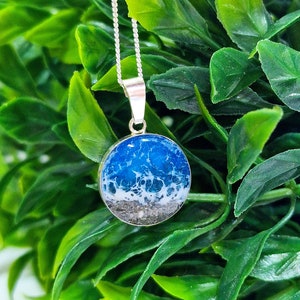 Pu&ograve; includere: Una collana con catena d'argento con un ciondolo rotondo che presenta un design a onda oceanica in resina blu e bianca. Il ciondolo ha uno strato di sabbia sul fondo.