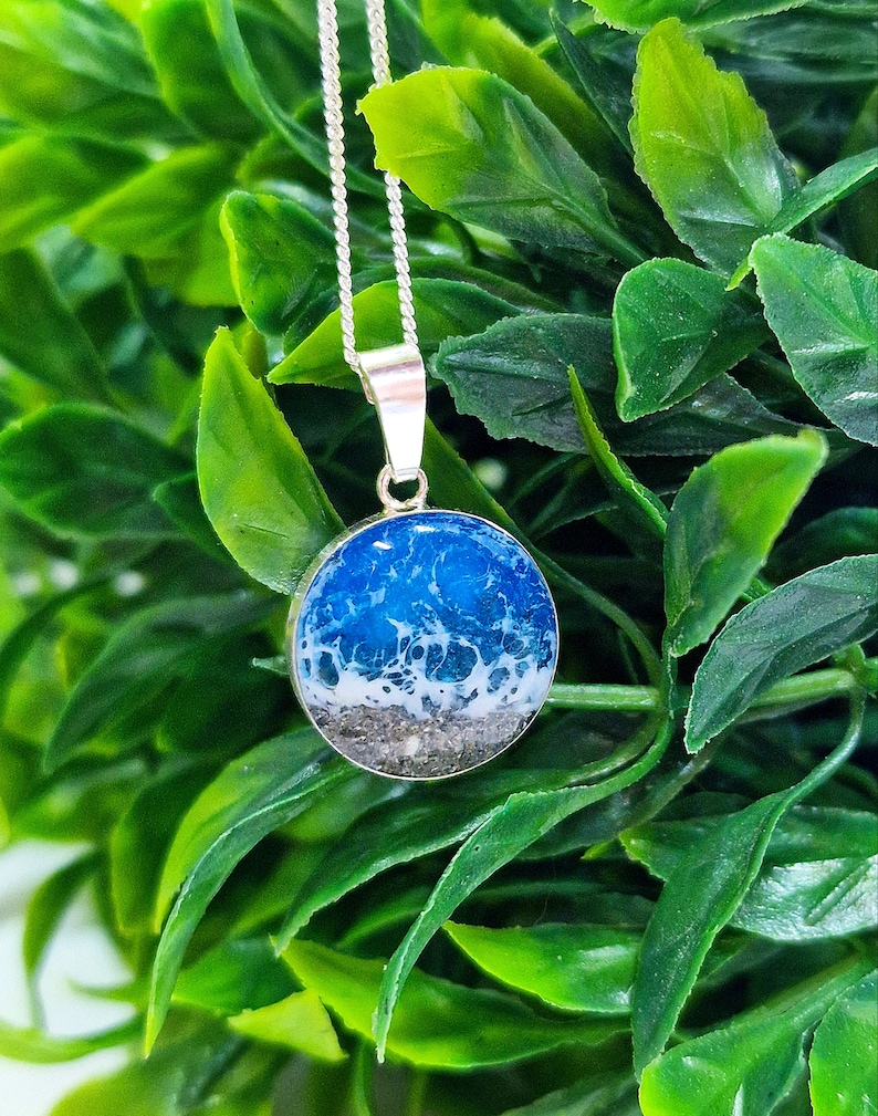 Pu&ograve; includere: Una collana con catena d'argento con un ciondolo rotondo che presenta un design a onda oceanica in resina blu e bianca. Il ciondolo ha uno strato di sabbia sul fondo.