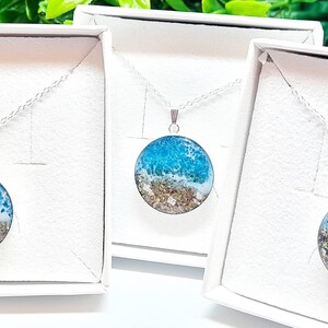 May include: Three silver necklaces with round pendants featuring a blue and white resin ocean scene with gold glitter. The necklaces are displayed in white boxes.