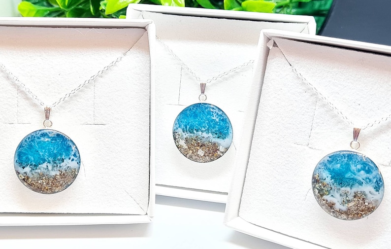 May include: Three silver necklaces with round pendants featuring a blue and white resin ocean scene with gold glitter. The necklaces are displayed in white boxes.