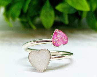 Può includere: Un anello argentato con due pietre a forma di cuore. Un cuore è di colore rosa chiaro, l'altro è bianco. L'anello ha un design avvolgente, con i cuori posizionati su lati opposti della fascia.
