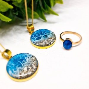 May include: Two gold necklaces with round blue and white resin pendants depicting ocean waves and a gold ring with a blue resin stone.