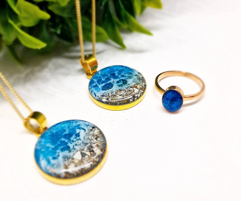 May include: Two gold necklaces with round blue and white resin pendants depicting ocean waves and a gold ring with a blue resin stone.