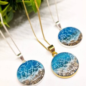 May include: Three necklaces with round pendants featuring a blue and white ocean scene with sand and shells. The pendants are on silver and gold chains.