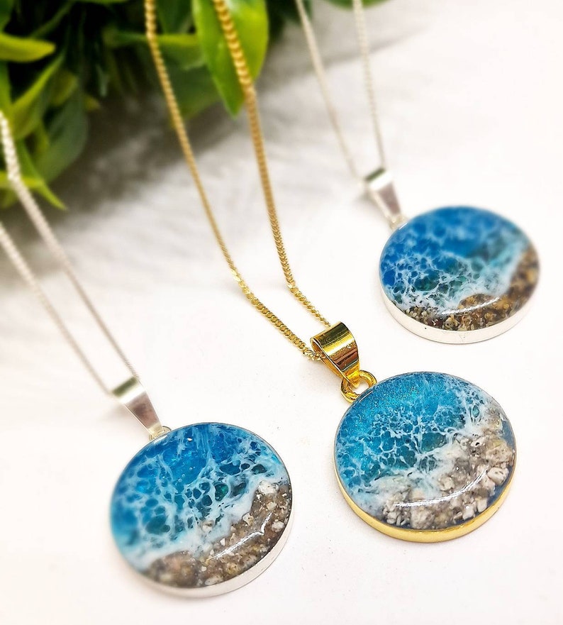 May include: Three necklaces with round pendants featuring a blue and white ocean scene with sand and shells. The pendants are on silver and gold chains.