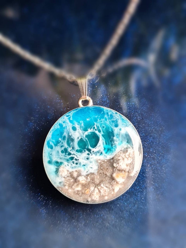 May include: A round resin pendant necklace featuring a blue and white ocean wave design with a brown sandy beach at the bottom. The pendant is on a silver chain.
