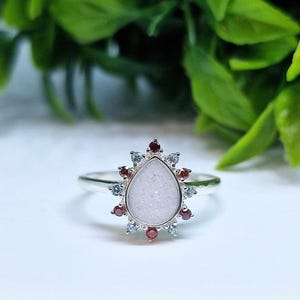 May include: A silver ring featuring a white teardrop-shaped stone encircled by small red and clear stones.