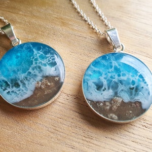 May include: Two silver necklaces with round pendants featuring a resin ocean scene. The pendants are filled with blue and white resin that resembles waves crashing on a sandy beach.