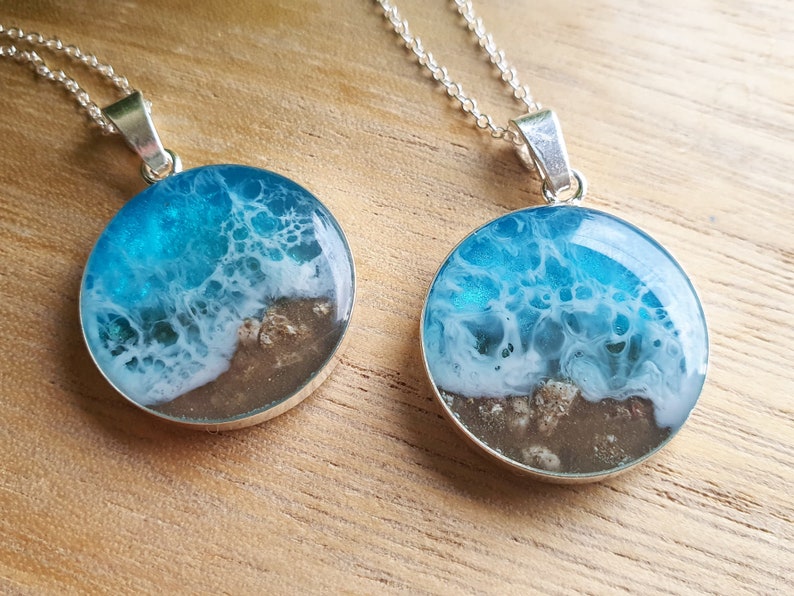 May include: Two silver necklaces with round pendants featuring a resin ocean scene. The pendants are filled with blue and white resin that resembles waves crashing on a sandy beach.