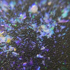 May include: Close-up of a dark surface covered in iridescent, multi-colored flakes. The flakes are primarily shades of blue and purple, with hints of green and yellow. The surface appears textured.