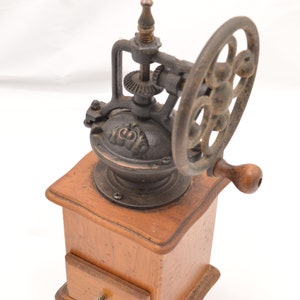 May include: Antique wooden coffee grinder with a cast iron mechanism and a crank handle. The grinder has a drawer for storing coffee beans.