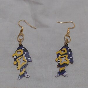 May include: A pair of gold fish-shaped earrings with purple, yellow, and white paint. The fish have a cutout design and white dots.