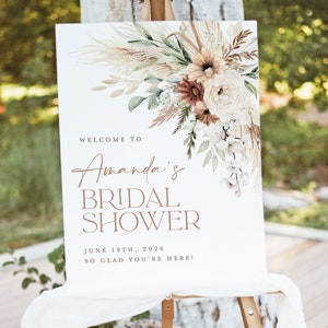 Op de afbeelding: Een wit bord op een ezel met bloemstukken en de tekst "Welkom bij Amanda's Bridal Shower" met de datum 19 juni 2026. Het bloemstuk bevat pampasgras, rozen en andere bloemen in neutrale tinten.