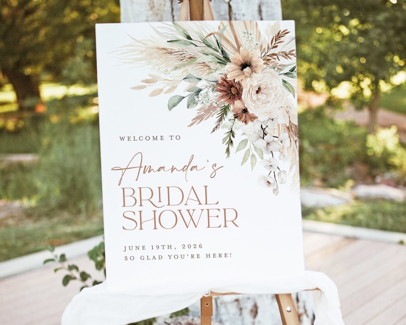 Op de afbeelding: Een wit bord op een ezel met bloemstukken en de tekst "Welkom bij Amanda's Bridal Shower" met de datum 19 juni 2026. Het bloemstuk bevat pampasgras, rozen en andere bloemen in neutrale tinten.