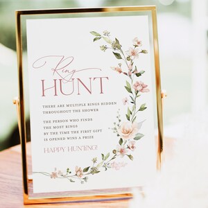 May include: A white sign with a floral border and the text "Ring Hunt" in pink. The sign says "There are multiple rings hidden throughout the shower. The person who finds the most rings by the time the first gift is opened wins a prize. Happy Hunting!"