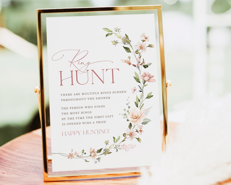 May include: A white sign with a floral border and the text "Ring Hunt" in pink. The sign says "There are multiple rings hidden throughout the shower. The person who finds the most rings by the time the first gift is opened wins a prize. Happy Hunting!"