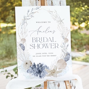 Pode incluir: Uma placa branca num cavalete, anunciando "Amelia's Bridal Shower". A placa apresenta um design em aquarela de conchas, estrelas do mar e corais em tons de azul, cinza e bege. A data é 19 de junho de 2056.