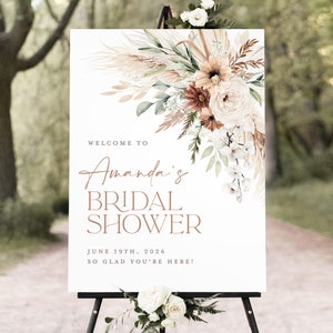Op de afbeelding: Een wit bord op een zwart schildersezel met bloemstukken. Het bord leest "Welcome to Amanda's Bridal Shower" met de datum 19 juni 2026 en de zin "So glad you're here!" in een bruin script.