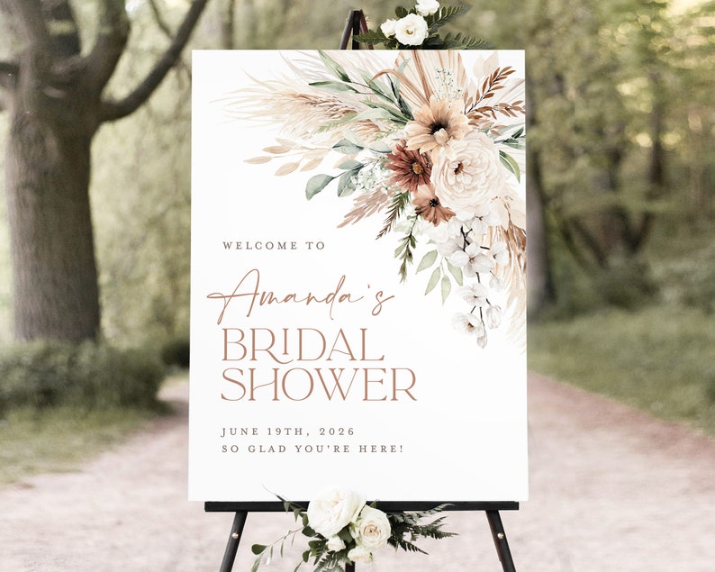 Op de afbeelding: Een wit bord op een zwart schildersezel met bloemstukken. Het bord leest "Welcome to Amanda's Bridal Shower" met de datum 19 juni 2026 en de zin "So glad you're here!" in een bruin script.