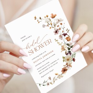 May include: A white bridal shower invitation with a floral wreath design in shades of orange, brown, and white. The text reads "PLEASE JOIN US FOR A bridal SHOWER celebrating bride-to-be OLIVIA JOHNSON SATURDAY, MAY 16TH AT 5:00PM STUDIO DE LA FLOR 2205 FARADAY AVE, CARLSBAD, CA RSVP TO NAME AT 532.783.226 Registered at Target"