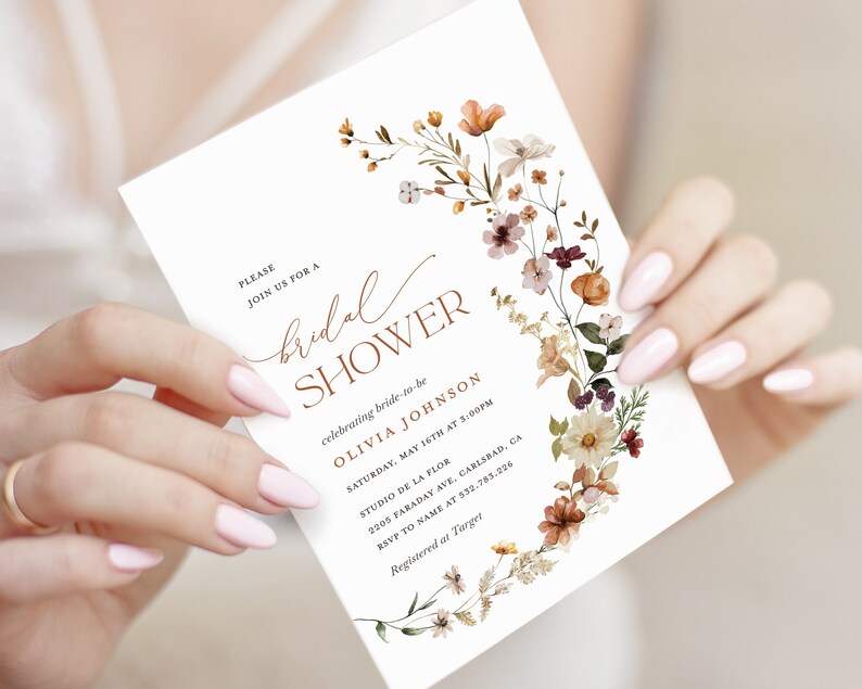 May include: A white bridal shower invitation with a floral wreath design in shades of orange, brown, and white. The text reads "PLEASE JOIN US FOR A bridal SHOWER celebrating bride-to-be OLIVIA JOHNSON SATURDAY, MAY 16TH AT 5:00PM STUDIO DE LA FLOR 2205 FARADAY AVE, CARLSBAD, CA RSVP TO NAME AT 532.783.226 Registered at Target"