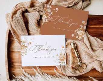 May include: Two "Thank you" cards with floral designs and the names "Amelia + Chandler" are displayed on a wooden tray. One card is white, the other is terracotta. A gold wax seal and stamp are also on the tray, along with a beige fabric.