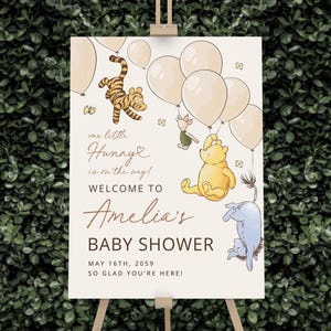 Pode incluir: Uma placa de boas-vindas para ch&aacute; de beb&ecirc; com personagens de Winnie the Pooh e bal&otilde;es. A placa inclui o texto "our little Hunny is on the way!" e "Welcome to Amelia's Baby Shower, May 16th, 2059". A placa est&aacute; em um cavalete de madeira.
