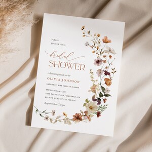 May include: A white bridal shower invitation with a floral wreath border in shades of orange, pink, and purple. The text reads "Please join us for a bridal shower celebrating bride-to-be Olivia Johnson Saturday, May 16th at 3:00PM Studio de la Flor 3205 Faraday Ave, Carlsbad, CA RSVP to name at 532-783-226 Registered at Target"