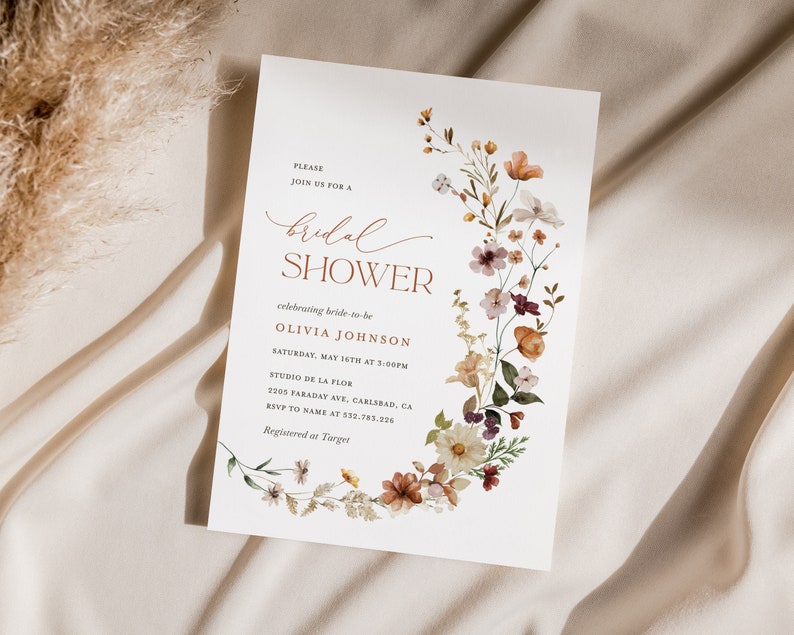 May include: A white bridal shower invitation with a floral wreath border in shades of orange, pink, and purple. The text reads "Please join us for a bridal shower celebrating bride-to-be Olivia Johnson Saturday, May 16th at 3:00PM Studio de la Flor 3205 Faraday Ave, Carlsbad, CA RSVP to name at 532-783-226 Registered at Target"