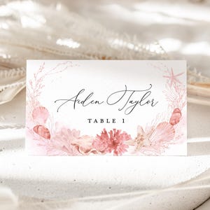 May include: A rectangular place card with the name "Aiden Taylor" in elegant script, and "TABLE 1" in a serif font. The card is decorated with a watercolor design of pink seashells, coral, and a starfish, creating a coastal theme.