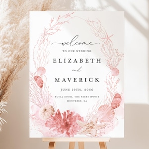 Puede incluir: Un letrero de bienvenida de boda con los nombres Elizabeth y Maverick, la fecha 19 de junio de 2056 y el lugar. El letrero está decorado con coral, conchas marinas y una estrella de mar en tonos rosa y blanco. Se muestra en un caballete de madera.