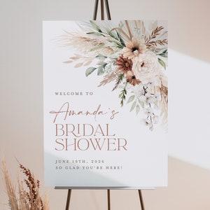 Op de afbeelding: Een wit bord op een ezel met de tekst "Welcome to Amanda's Bridal Shower" in bruine script- en blokletters. Het bord is versierd met een bloemstuk in neutrale tinten.