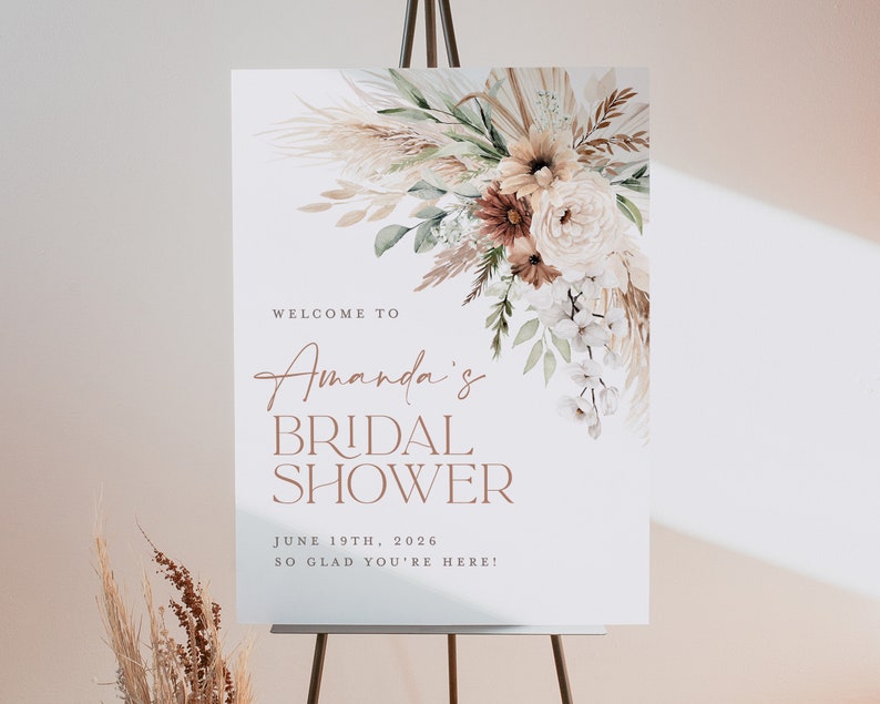Op de afbeelding: Een wit bord op een ezel met de tekst "Welcome to Amanda's Bridal Shower" in bruine script- en blokletters. Het bord is versierd met een bloemstuk in neutrale tinten.
